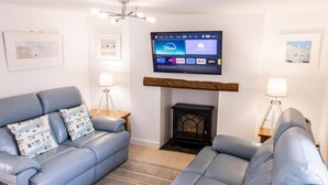 A living room with a fireplace, two blue sofas, a flat-screen TV mounted above the fireplace, and framed pictures on the walls. - 2 Bed in Shaldon (oc-t36353) (Shaldon)