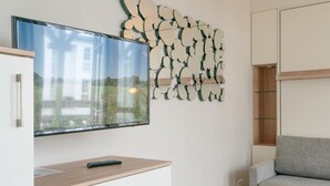 A modern living room with a flat-screen TV mounted on the wall, a wooden cabinet with a remote control, a grey sofa, and a decorative wall art piece. - Perfekt fur Zwei - Moderne Fewo mit Balkon in Sellin (Sellin)