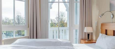 Quarto com cama, mesa de cabeceira com abajur, janela com cortinas e vista para árvores lá fora.