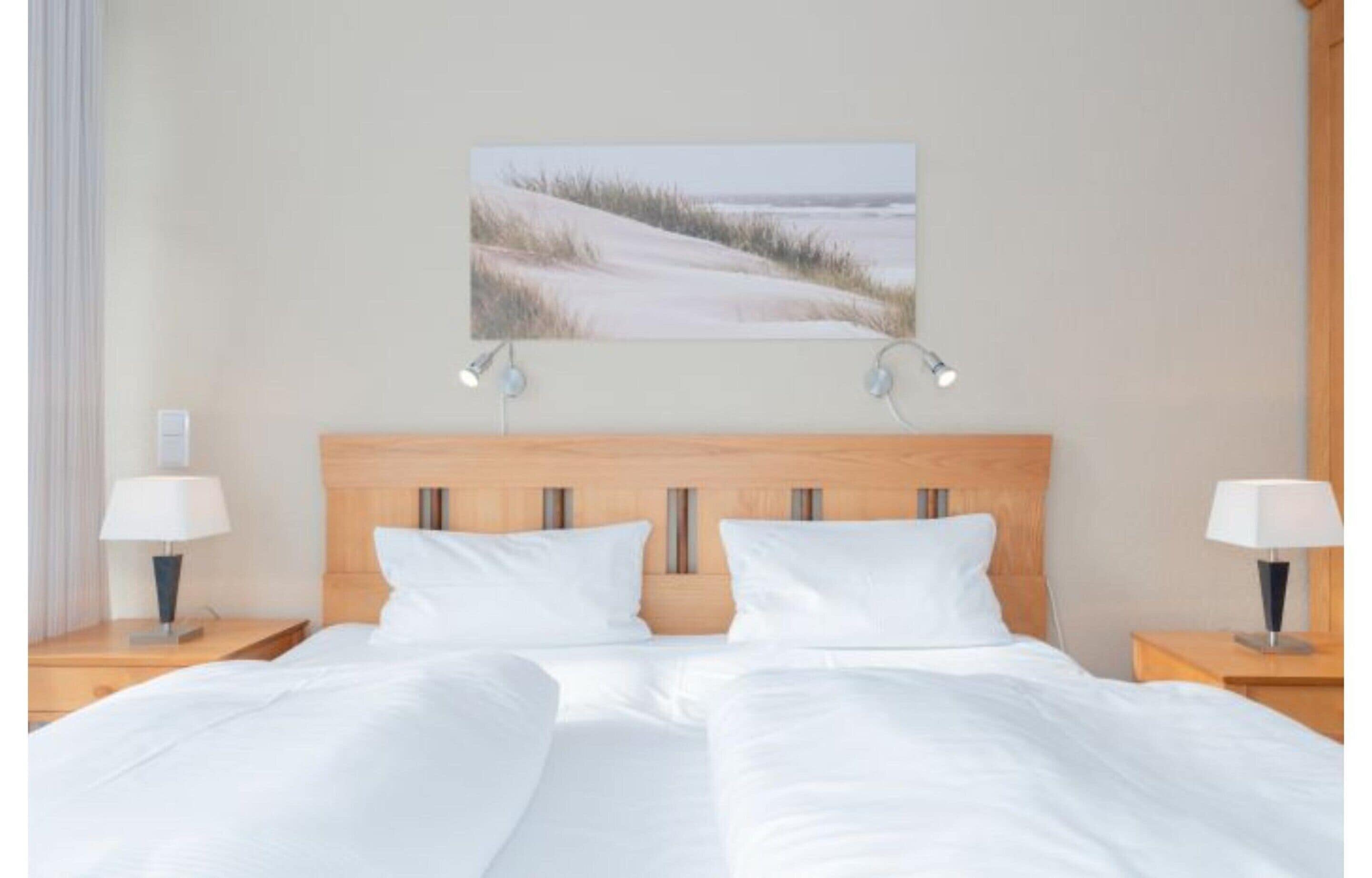 Ein Doppelbett mit weißer Bettwäsche, ein Holz-Kopfteil, Nachttischlampen und ein an der Wand befestigtes Bild von einem Strand.