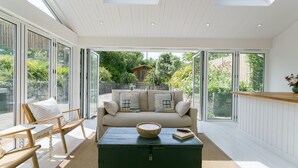 A modern living room with a sofa, coffee table, and chairs, large windows, and a view of a garden and house.