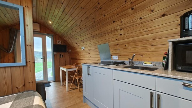 A compact kitchen with wooden walls, a dining area, and a view through a glass door. - 1 Bed in Tyn-y-Groes (oc-g36300) (Ty'n-y-groes)