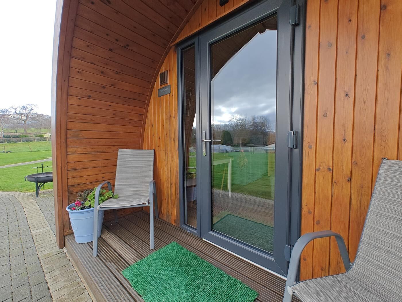 Une structure extérieure en bois avec une porte coulissante en verre, un patio couvert avec deux chaises et un pot de fleurs, et un tapis vert sur le sol.