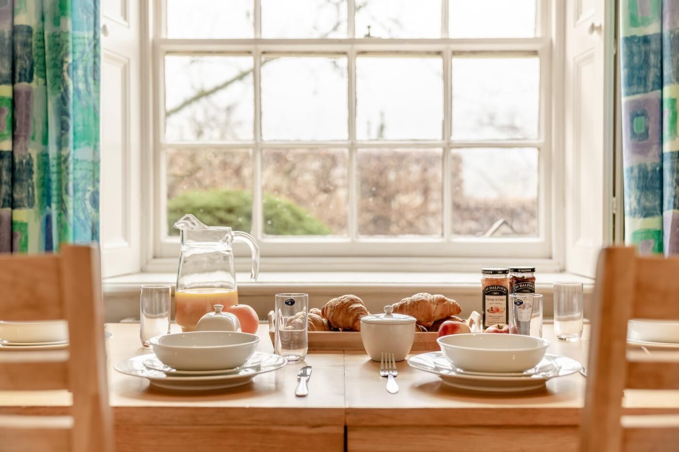 A breakfast table set with plates, a pitcher of orange juice, croissants, and a jar of jam.