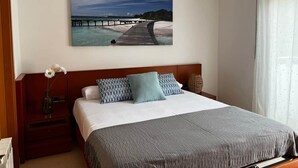 A hotel room with a bed, a nightstand with a vase, a painting of a pier, and a window with curtains. - La Mota Beach House with Pool & BBQ (Cubelles)