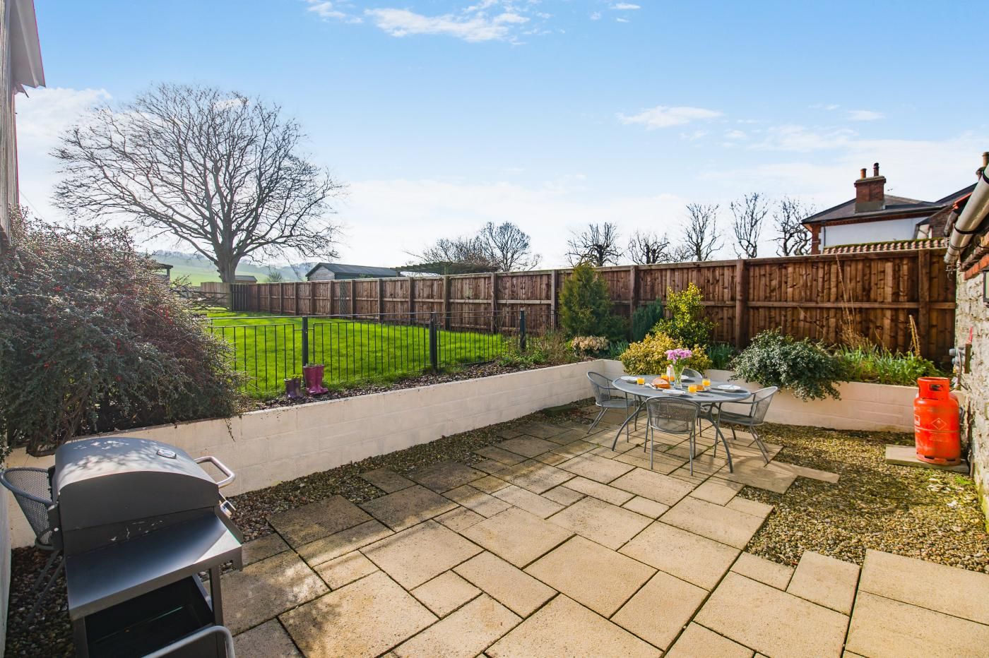 A patio with a grill, a table with chairs, and a view of a grassy yard and wooden fence.