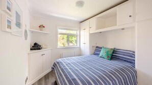 Ein Schlafzimmer mit einem Bett, einem Fenster, einer Heizung, einem Ablagefach mit Handtüchern und einem an der Wand befestigten Schrank mit einer Vase.