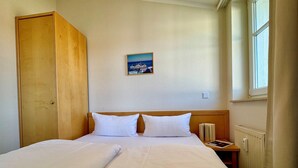 Quarto de hotel com cama de casal, guarda-roupa de madeira, mesa de cabeceira com um livro, uma pequena mesa e uma janela com cortinas.