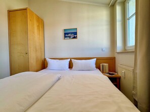 A hotel room with a double bed, a wooden wardrobe, a nightstand with a book, a small table, and a window with curtains. - Ferienwohnung Ostseenah mit Balkon und Ahoi-rabatt (Sellin)