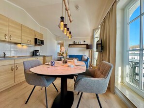 A modern kitchen with wooden cabinets, a dining table with chairs, a hanging light fixture, and a large window with curtains. - Ferienwohnung Ostseenah mit Balkon und Ahoi-rabatt (Sellin)