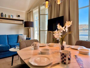 A modern living room with a blue sofa, a dining table set with plates and a coffee cup, a flat-screen TV, and a large window with curtains. - Ferienwohnung Ostseenah mit Balkon und Ahoi-rabatt (Sellin)