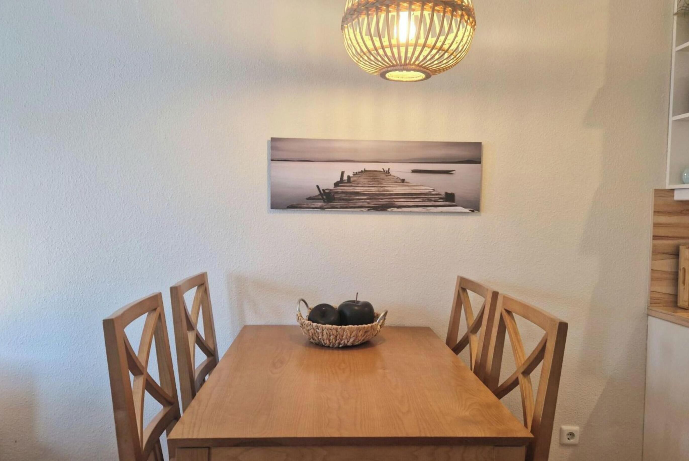 A dining table with chairs, a basket of apples, a wall-mounted picture, and a hanging light fixture.
