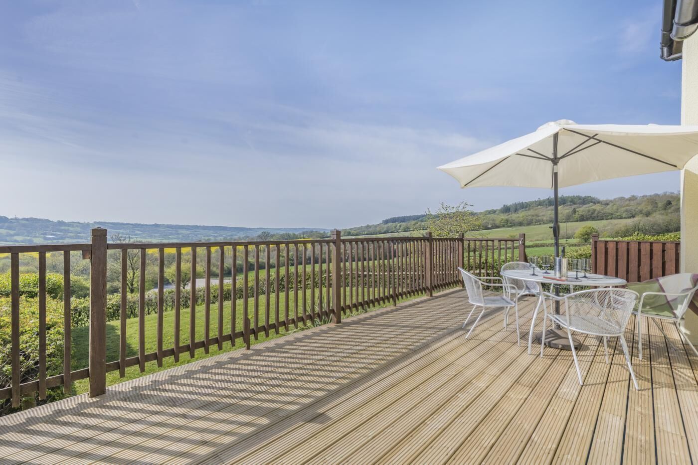 A wooden deck with a table and chairs, a large umbrella, and a view of a green landscape.