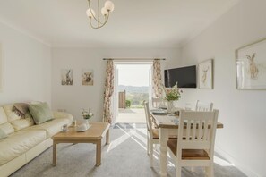 Ein Wohnzimmer mit einer Couch, einem Holztisch und einem Esstisch mit Stühlen. Ein Fernseher ist an der Wand befestigt und es gibt ein Fenster mit Vorhängen.
