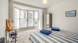 Ein Schlafzimmer mit einem großen Erkerfenster, einem Bett mit gestreifter Bettwäsche, einem Kleiderschrank und einem Schreibtisch mit Lampe.