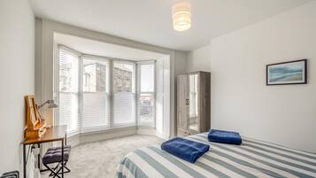 A bedroom with a large bay window, a bed with striped bedding, a wardrobe, and a desk with a lamp.