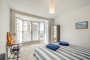 Ein Schlafzimmer mit einem großen Erkerfenster, einem Bett mit gestreifter Bettwäsche, einem Kleiderschrank und einem Schreibtisch mit Lampe.