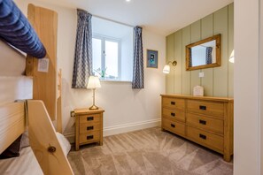 A bedroom with a bed, a nightstand with a lamp, a dresser with drawers, a window with curtains, and a mirror on the wall. - 2 Bed in Padstow (oc-g34917) (Trevone)