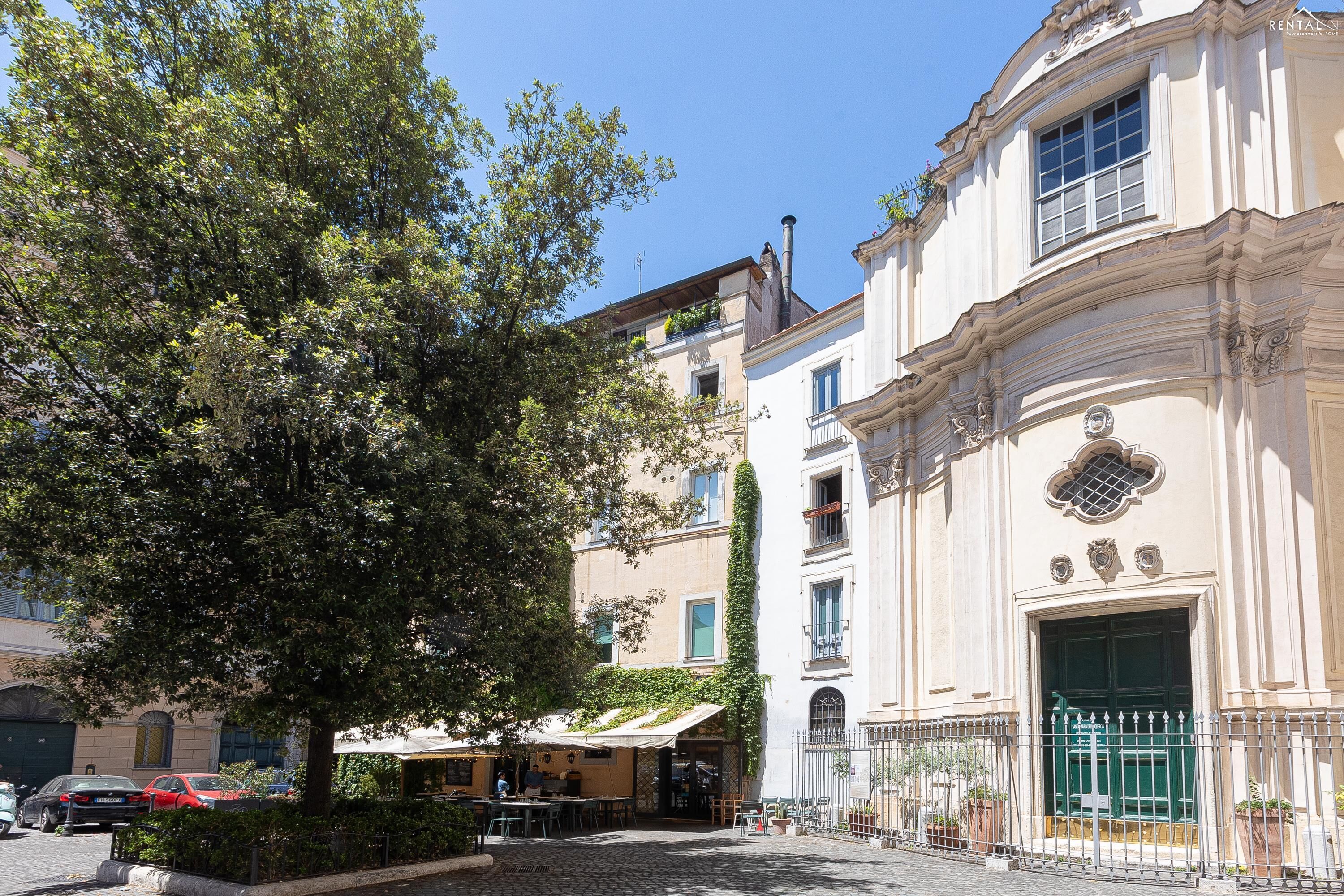 A cobblestone street with a building featuring a large tree, outdoor seating, and a green gate.