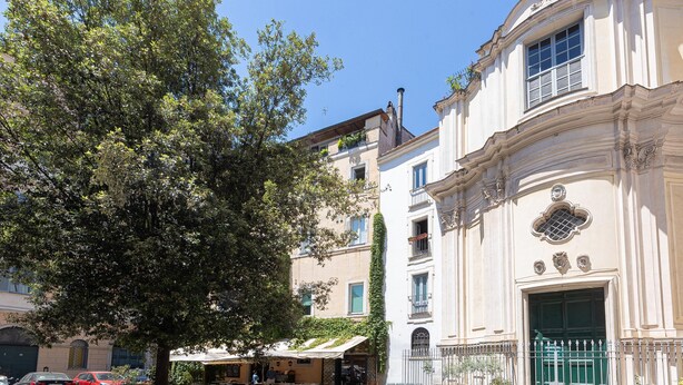 A cobblestone street with a building featuring a large tree, outdoor seating, and a green gate. - Red Hat Home by Campo de Fiori (Rome)