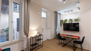 A modern living room with a TV, a dining table with chairs, a small side table with a lamp, and a large window with curtains. - Red Hat Home by Campo de Fiori (Rome)
