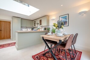 A modern kitchen with a central island, a dining area with a table set for four, and a red patterned rug on the floor. - 2 Bed in Padstow (oc-f34918) (Trevone)
