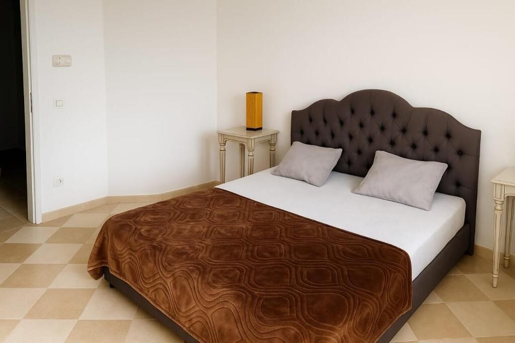 Une chambre à coucher avec une tête de lit capitonnée, une couvre-lit à motifs, deux oreillers et une table de chevet avec une lampe.