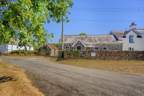 Un chemin de campagne mène à une maison en pierre dotée d’un toit à pignon, entourée d’herbe sèche et d’un mur de pierres.