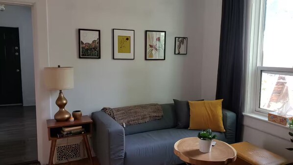 Ett vardagsrum med en soffa, en lampa, ett litet bord och ett fönster med gardiner.