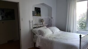 Ett sovrum med en sängram i metall, vita sängkläder, ett fönster med gardiner och ett konstverk som hänger på väggen.