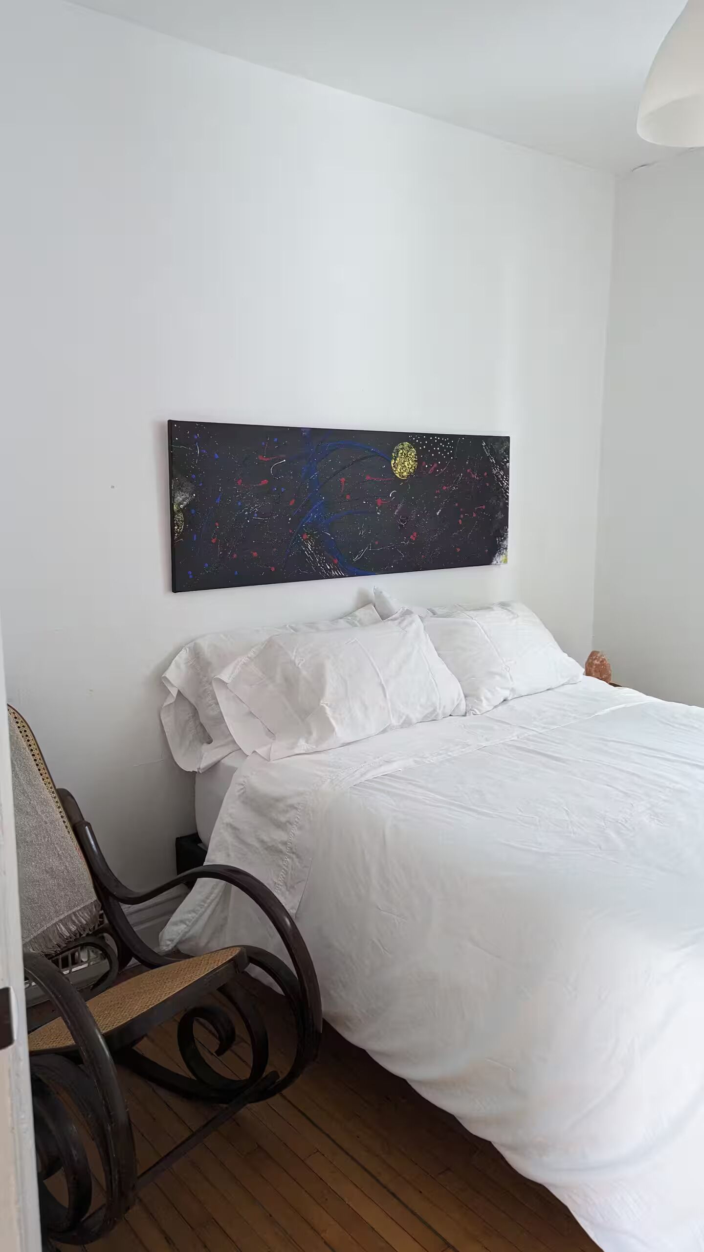 Een slaapkamer met een bed, witte beddengoed, kunstwerk van een nachthemel aan de muur en een houten vloer.