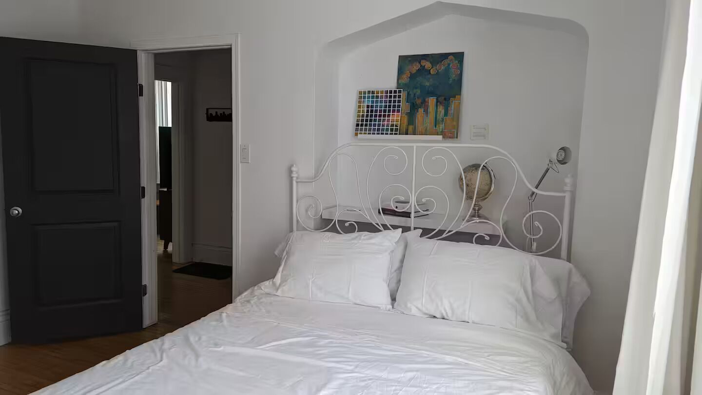 Un dormitorio con una cama blanca, una puerta, un pasillo y un cuadro en la pared.