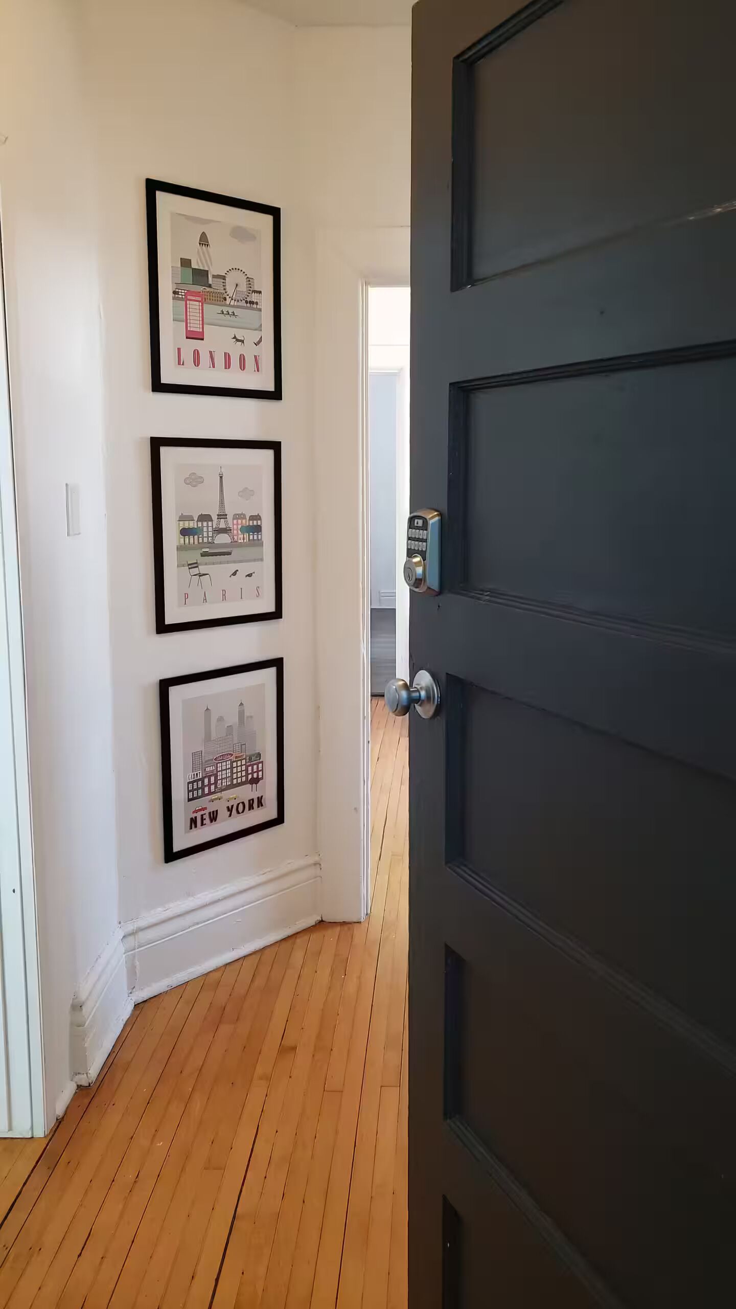 Un pasillo con cuadros de paisajes urbanos en la pared, suelo de madera y una puerta oscura.