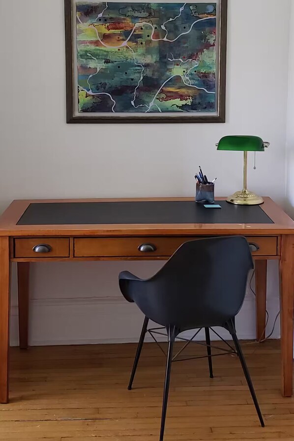 Un bureau en bois avec deux tiroirs, une chaise noire, un tableau abstrait encadré et une lampe de bureau verte.