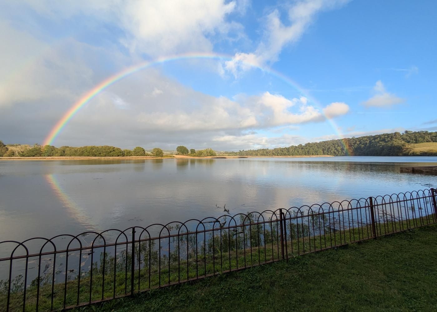A calm lake with a rainbow, a metal fence, and trees in the background.