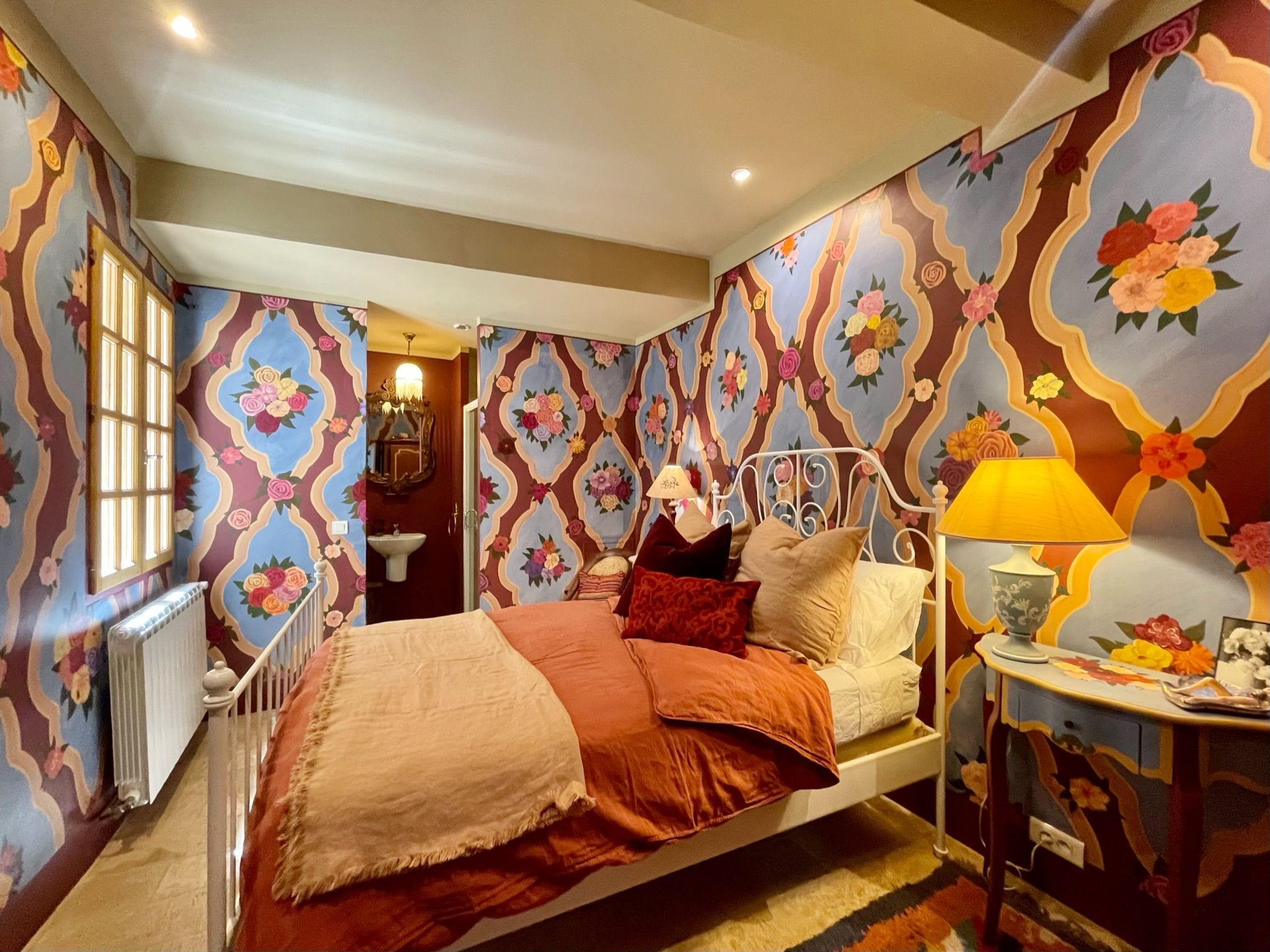 Un dormitorio con cama, mesita de noche con lámpara y papel tapiz floral.