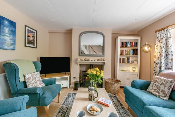 Ein Wohnzimmer mit türkisfarbenen Sofas, einem Flachbildfernseher, einem Kamin, einem Bücherregal und einem Fenster mit floralem Vorhang.