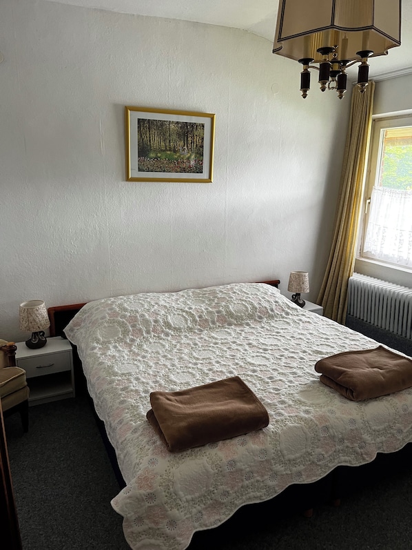 Un dormitorio con cama, mesita de noche, lámpara, un cuadro en la pared y una ventana con cortinas.