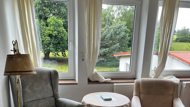 Ein Zimmer mit zwei Sesseln, einem kleinen Tisch, einer Stehlampe und großen Fenstern mit Vorhängen.