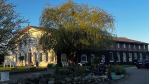Ein Gebäude mit einer Stein-Fassade, ein großer Baum davor und ein gepflasterter Bereich mit Gartenmöbeln.