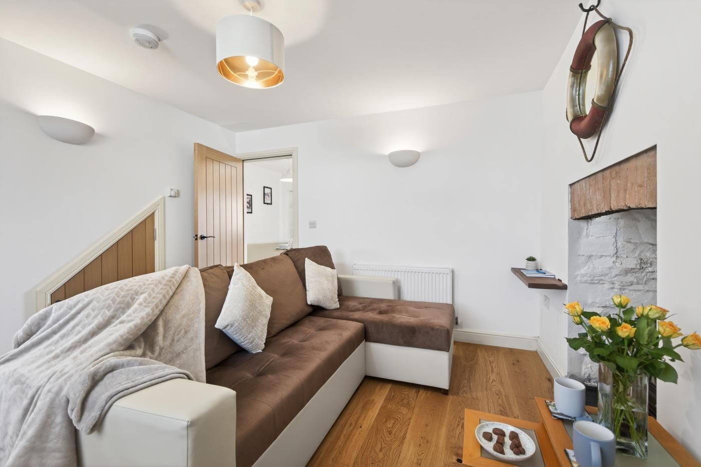 A modern living room with a brown sectional sofa, a white armchair, a wooden floor, a white radiator, and a wall-mounted life preserver.