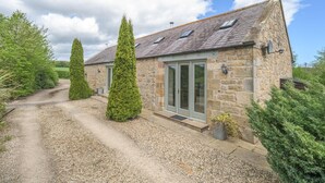 A stone house with a gravel driveway, tall coniferous trees, and a clear sky. - 2 Bed in Christon Bank (oc-coq128) (Christon Bank)