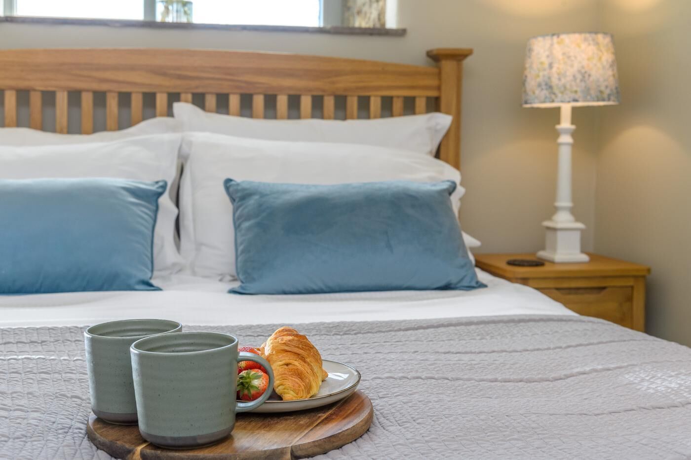 A neatly made bed with blue pillows, a bedside table with a lamp, and a tray with a croissant and a cup.