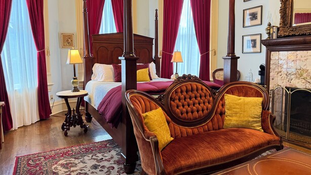 Una cama con dosel y cabecera de madera, una silla tapizada en rojo y amarillo, una alfombra estampada y una chimenea con repisa de azulejos.