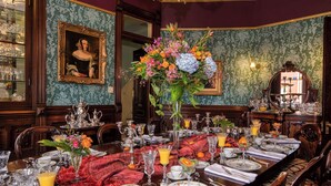 Comedor formal con una larga mesa dispuesta para una comida, rodeado de muebles ornamentados y un gran centro de mesa floral.