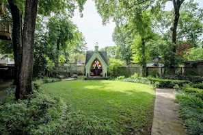 A small, green-roofed structure with a pointed arch and a stained glass window, surrounded by a well-maintained garden and a wooden fence.