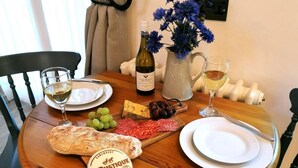 Een houten tafel met een fles wijn, een karaf water, een glas witte wijn, een brood, een kaasplank, druiven en een pot mosterd.