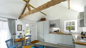 Een kleine keuken met witte kastjes, een magnetron en een spoelbak. Er is een eethoek met een tafel en stoelen, en een bank met kleurrijke kussens.