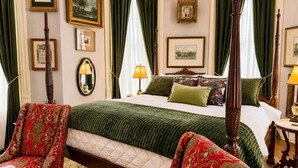 Une chambre à coucher avec un lit à baldaquin, des rideaux verts, des tableaux encadrés aux murs et un fauteuil rouge à motifs.
