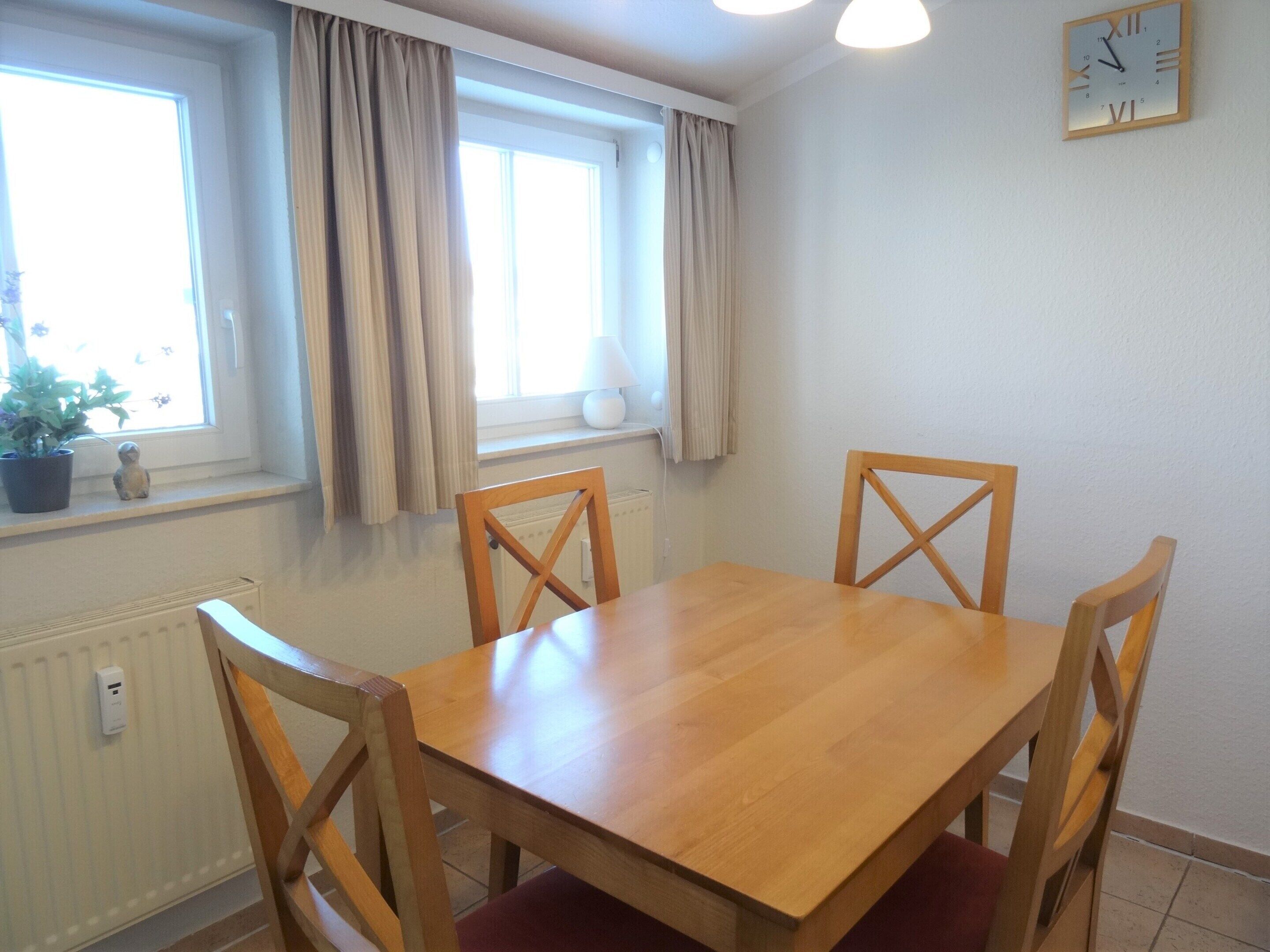 Ein Esszimmer mit Holztisch und vier Stühlen, einem fenster mit Vorhängen, einem Heizkörper und einer Wanduhr.